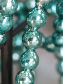 A close-up of hanging glass beads in a metallic teal-green color, showing reflective surfaces and a soft blurred background.