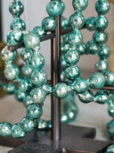 A close-up of a decorative glass bead sculpture or garland made of glossy teal-blue marbled glass beads hanging from a dark metal stand.