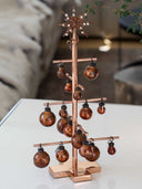 A copper-colored metal Christmas tree display stand with multiple branches holding round copper-toned ornaments, shown on a white tabletop in a softly blurred indoor setting.