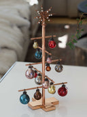 A copper-colored decorative Christmas tree stand with multiple hanging glass ornament bulbs in assorted colors, displayed on a white tabletop with a softly blurred indoor background.