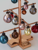 A copper-colored metal Christmas tree display stand holding multiple vintage-style glass ornaments in metallic colors including blue, red, gold, bronze, silver, and pink, photographed against a light neutral background.