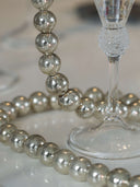A close-up of a silver-beaded decorative garland draped around a clear glass stemware base on a light reflective surface.