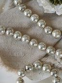 A close-up of shiny silver glass bead garland draped across a beige linen fabric with fringe, with a white flower in the corner.