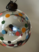 A large, spherical, highly reflective silver Christmas ornament decorated with colorful polka dots in orange, green, blue, pink, black, and white. The ornament is shown hanging against a plain beige wall, with its reflection visible on its shiny surface.