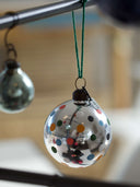A clear glass Christmas ornament decorated with colorful polka dots, hanging from a green string from a metal rod. Another silver ornament is blurred in the background.
